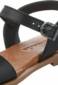 Black flat sandal with a leather upper, ankle strap, and silver buckle. Brown insole with subtle logos, low-profile tan outsole.