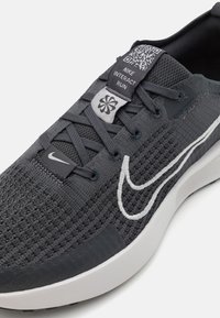 Chaussures de course Nike Interact en tissu tricoté gris foncé. Caractéristiques : tissu texturé, logo swoosh blanc, et code QR sur la languette. Accents de semelle blanche.