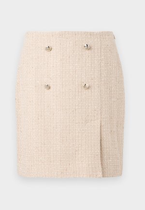 Gonna beige strutturata con un motivo intrecciato, caratterizzata da due bottoni argentati prominenti sul davanti e un orlo leggermente asimmetrico.