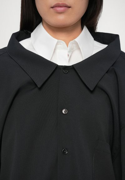 Detailní záběr na osobu oblečenou v vrstveném outfitu s bílou košilí s límcem pod černým kabátem na knoflíky s širokými klopami.