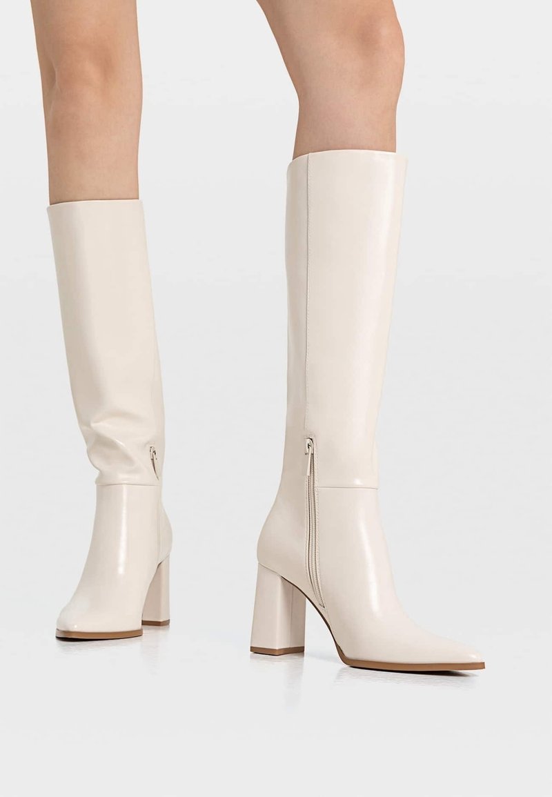Bottes blanches brillantes à hauteur de genou avec un bout pointu et un talon épais. Elles disposent d'une fermeture éclair sur le côté et d'une texture en cuir lisse.
