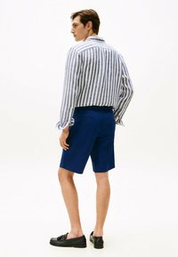 Gestreepte lange mouwen overhemd in wit en blauw, gecombineerd met effen blauwe shorts en zwarte instappers, gezien van achteren.