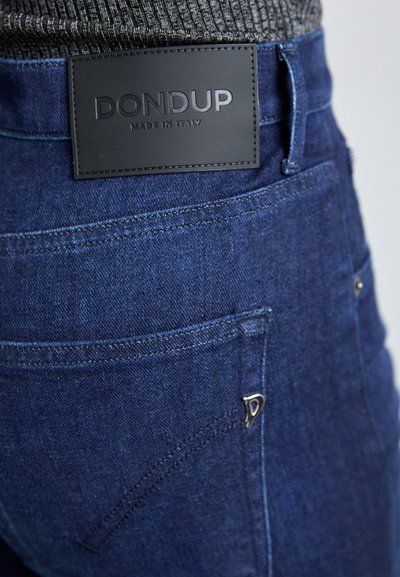Jeans de mezclilla azul oscuro con una textura suave. Presentan una etiqueta de cuero negro con la inscripción "DON DUP MADE IN ITALY" y un acento de hardware plateado en el bolsillo.