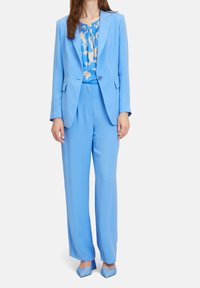 Kvinna klädd i en ljusblå kostym med en blazer med en knapp, matchande vida byxor och en blus med blått blommönster, i kombination med blå klackar.