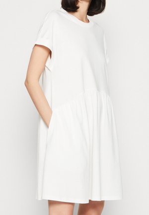 Frau trägt ein kurzärmeliges, locker sitzendes weißes Kleid mit geraffter Taille und Taschen, vor einem schlichten weißen Hintergrund.