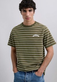Camiseta a rayas caqui con rayas horizontales blancas, cuello redondo acanalado y pequeño logo de "scalpers" en el pecho. Llevada con vaqueros azules.