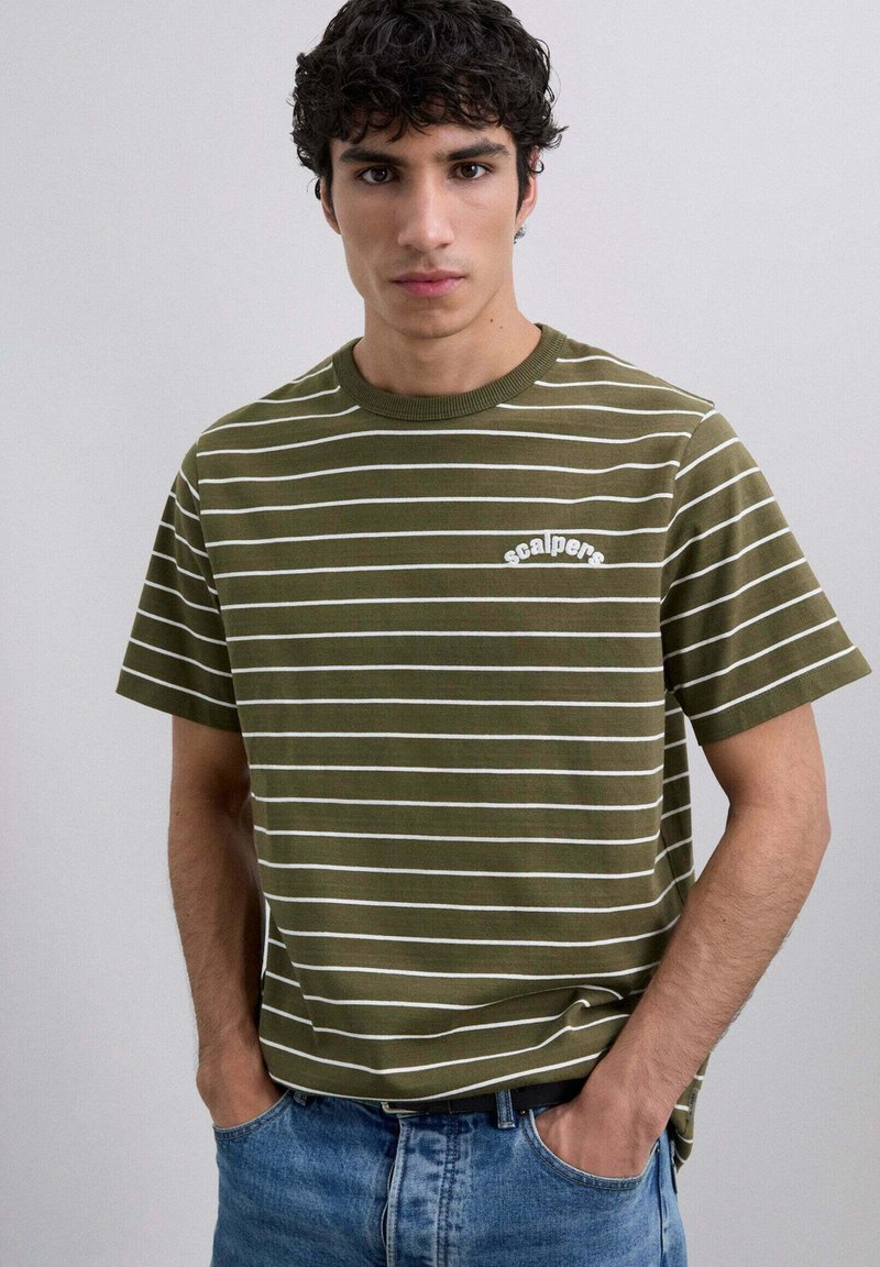 Camiseta a rayas caqui con rayas horizontales blancas, cuello redondo acanalado y pequeño logo de "scalpers" en el pecho. Llevada con vaqueros azules.