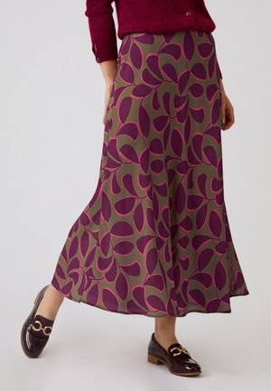 Mujer que lleva una falda larga verde oliva con grandes patrones de hojas moradas combinada con mocasines marrón oscuro brillantes con detalles dorados.
