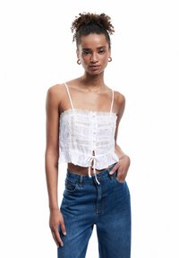 Top corto bianco con bordino in pizzo, spalline regolabili, chiusura frontale con bottoni e orlo con cordino. Abbinato a jeans blu a vita alta.