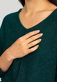 Femme portant un haut vert foncé scintillant à col en V, une main posée sur sa poitrine, montrant des ongles soigneusement manucurés dans une teinte nude.