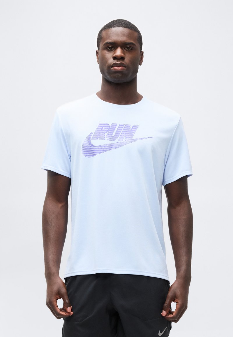 Άνδρας που φοράει ανοιχτό μπλε κοντομάνικο πουκάμισο με μωβ λέξη "RUN" και λογότυπο σβούρας, σε συνδυασμό με μαύρο σορτς, στέκεται μπροστά σε απλό φόντο.