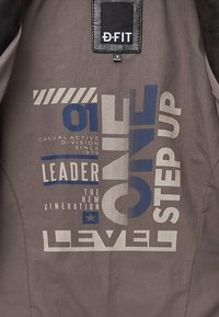 Donkergrijze stof met blauwe en witte tekst die leest: "LEIDER DE NIEUWE GENERATIE CASUAL ACTIEVE DIVISIE SINDS 1979 ÉÉN CHIQUE STAP OP NIVEAU."