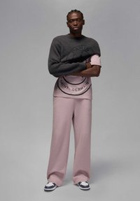 Mies seisoo käsivarret ristissä pukeutuneena vaaleanpunaiseen Saint-Germain-verryttelypukuun, mustavalkoisiin lenkkareihin ja tummanharmaa neule harteillaan.