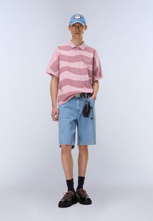 Junger Mann, der ein rosa gestreiftes Poloshirt, Jeansshorts, schwarze Schuhe mit pinken Schnürsenkeln, schwarze Socken und eine blaue Kappe mit einem Aufnäher trägt.