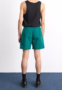 Zelené šortky s elastickým pasem, rovnou přední částí a bočními akcenty, kombinované s černým topem bez rukávů a tmavými ponožkami. Černé boty.