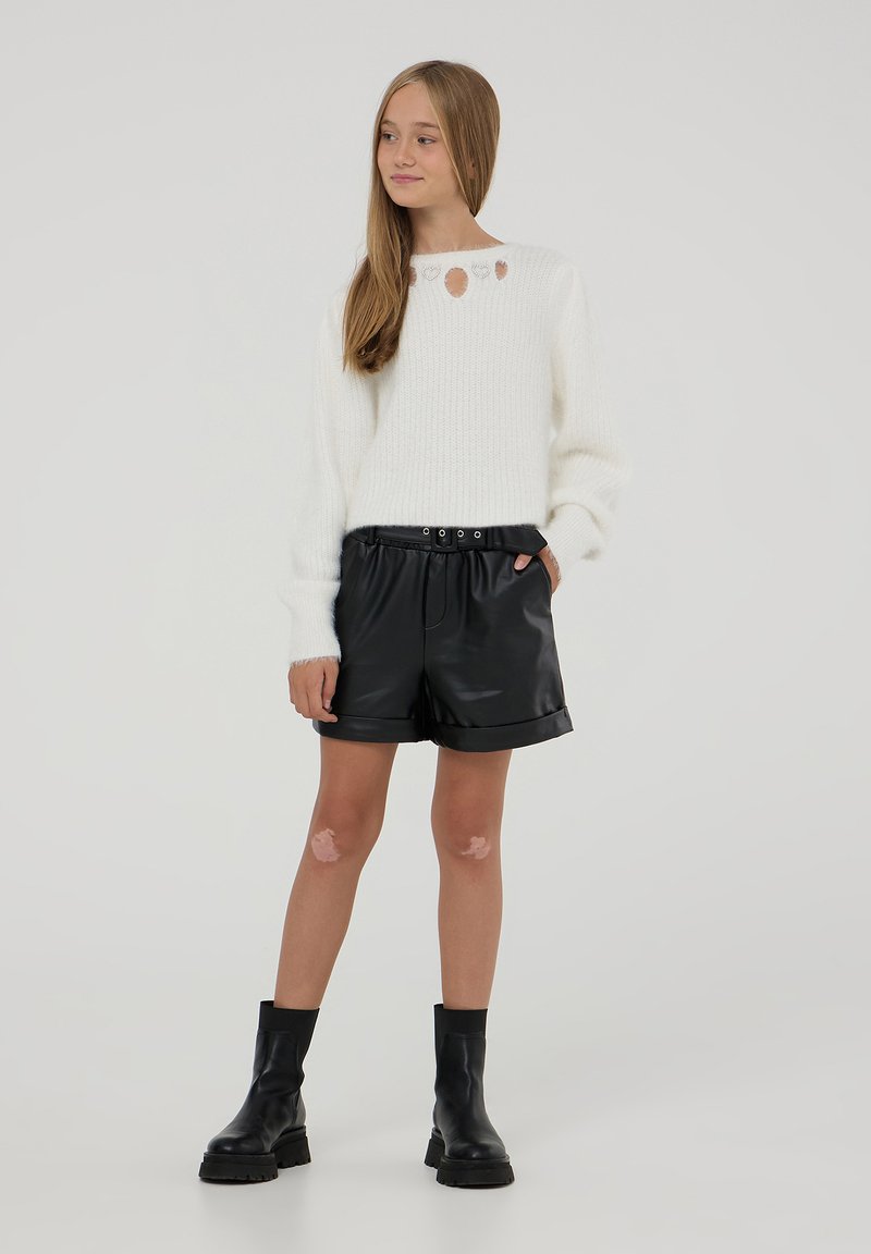 Maglione bianco lavorato a maglia con scollatura rotonda e fori decorativi, abbinato a pantaloni corte in finta pelle nera e stivaletti neri alla caviglia.