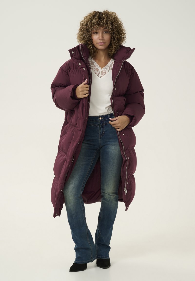 Lange, donkerviolette puffy coat met een hoge kraag, voorzien van drukknoopsluiting, gequilt textuur en een volle rits; gecombineerd met blauwe spijkerbroek en zwarte hakken.