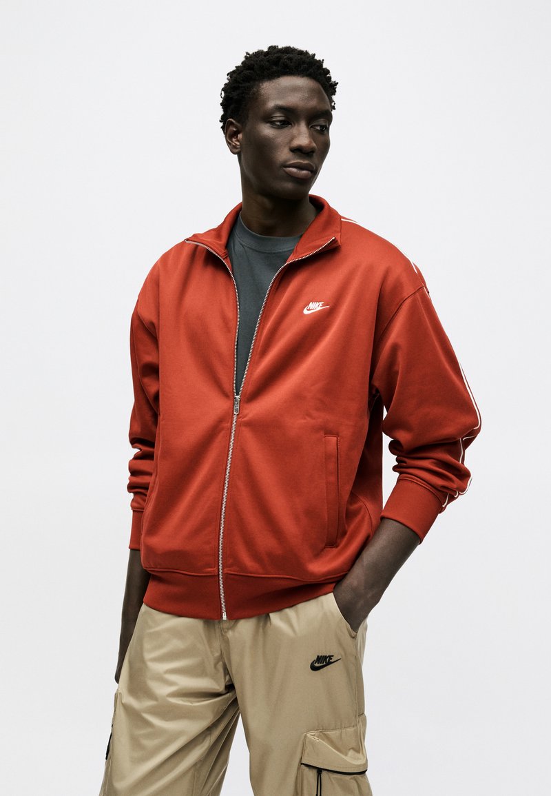 Uomo con capelli corti e ricci che indossa una giacca arancione Nike con zip sopra una maglietta scura e pantaloni cargo beige Nike, in piedi con una mano in tasca.