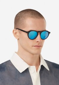 Kulaté sluneční brýle s černými obroučkami a modrými zrcadlovými čočkami, vyznačující se unikátním úhlovým designem. Model má na sobě námořnicky modrou a bílou košili s límečkem.