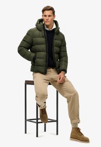 Giacca puffer verde scuro con cappuccio, texture trapuntata, indossata sopra un maglione blu navy a trecce. Pantaloni beige chiaro e stivali marroni con lacci completano il look.