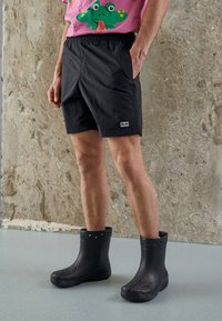 Botas pretas impermeáveis com superfície texturizada, acompanhadas de calções pretos com cintura elástica e um pequeno detalhe de logo.