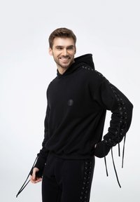 Czarna bluza z kapturem z sznurowaniami na rękawach, miękka tkanina, luźny krój i okrągłe logo z przodu. W połączeniu z pasującymi spodniami.