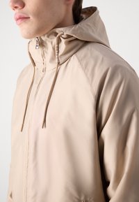 Ljusbeige jacka gjord av ett slätt, vattentätt material. Har en dragsko i huvan, dragkedjeslutning och subtilt präglad logodetalj.