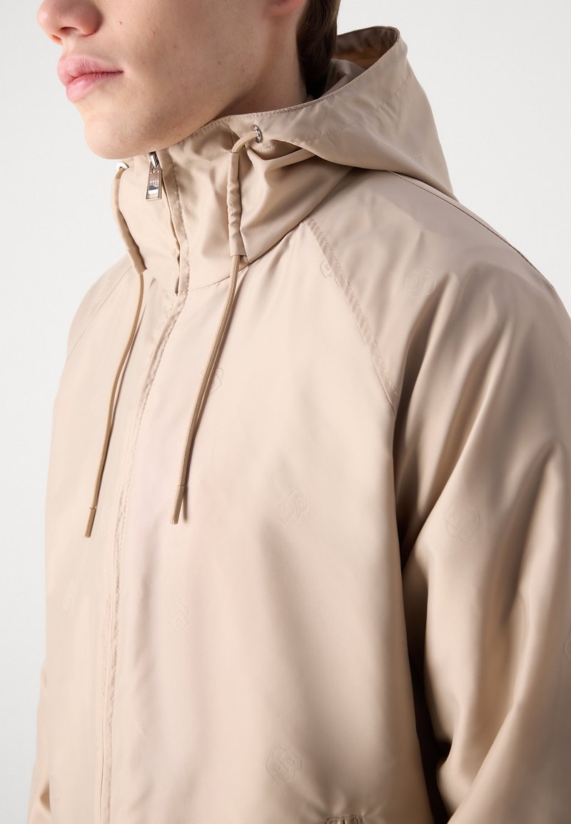 Veste beige clair en matériau lisse et imperméable. Dotée d'une capuche avec cordon de serrage, d'une fermeture éclair et d'un discret logo en relief.