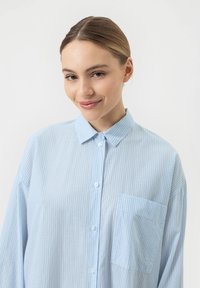 Chemise bleu clair à rayures, avec un col, une poche avant et des manches longues. Le tissu semble doux et respirant.