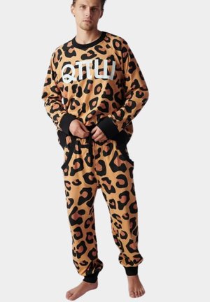 Mężczyzna w bluzie w panterkę i dopasowanych spodniach dresowych z czarnymi mankietami oraz białym napisem na piersi, stojący boso.