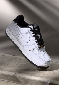 Hvit Nike Air Force 1-sneaker med svart såle, svarte snøringer, svart krave og liten gull Nike-svulst nær tåen, utstilt på grå overflate.