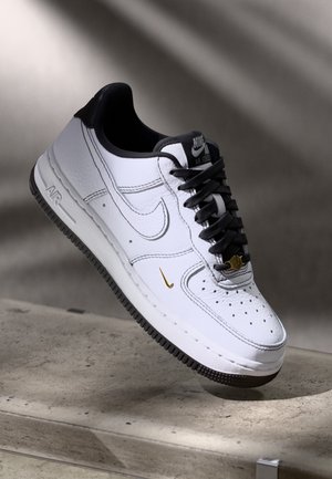 Zapatilla blanca Nike Air Force 1 con suela negra, cordones negros, cuello negro y pequeño logo dorado de Nike cerca de la punta, exhibida sobre una superficie gris.