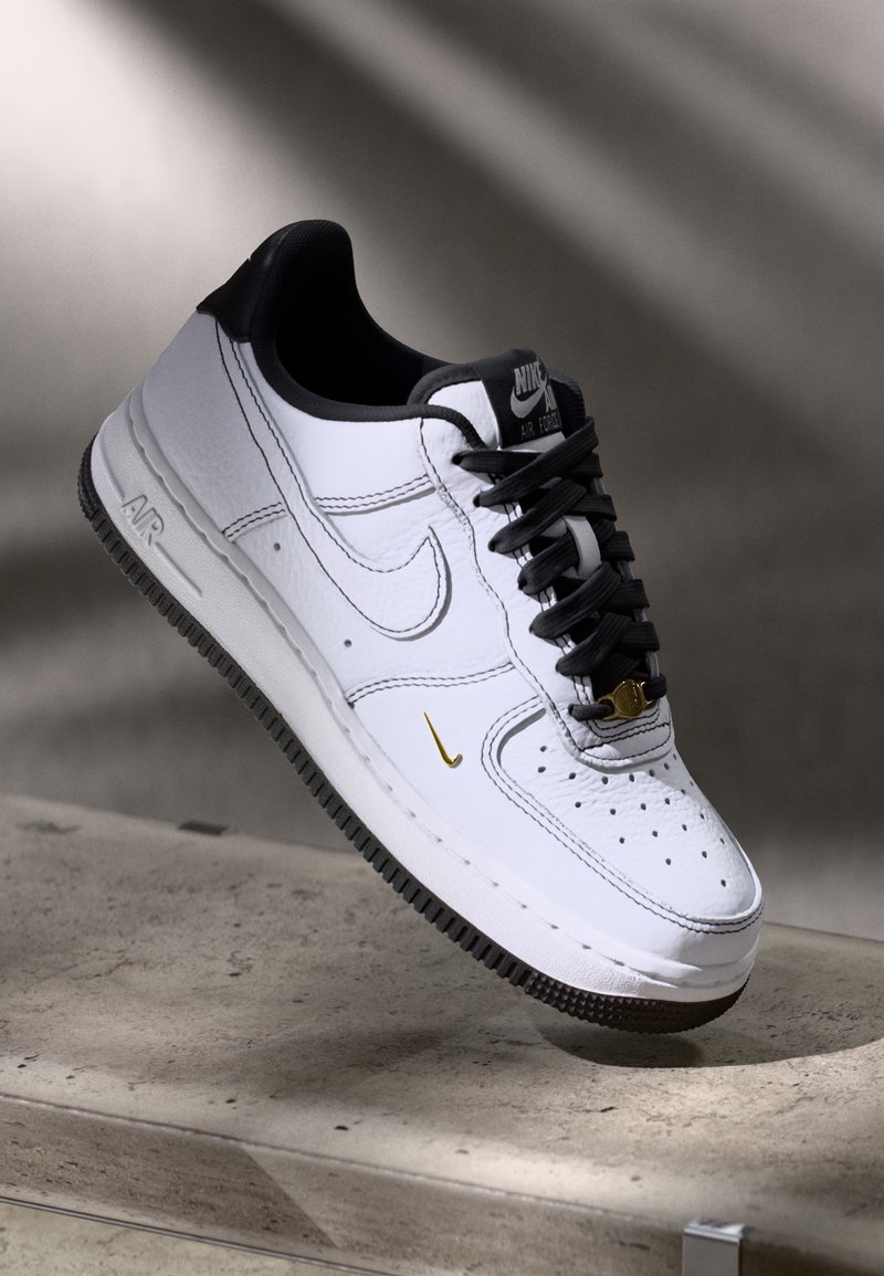 Hvit Nike Air Force 1-sneaker med svart såle, svarte snøringer, svart krave og liten gull Nike-svulst nær tåen, utstilt på grå overflate.