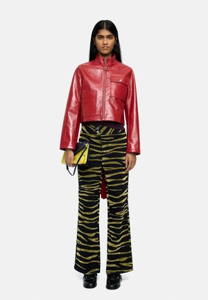 Kobieta stojąca, ubrana w błyszczącą czerwoną kurtkę na suwak, czarne spodnie w żółte paski tygrysie, czarne buty, trzymająca żółtą i niebieską torebkę.