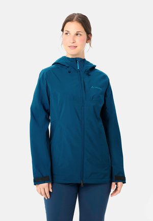 Donna che indossa una giacca impermeabile con cappuccio color teal, con zip frontale e polsini regolabili, che guarda leggermente di lato.