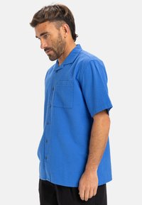 Camisa de botones de manga corta en tejido azul brillante texturizado, con un único bolsillo en el pecho y un corte holgado. Botones y cuello negros.