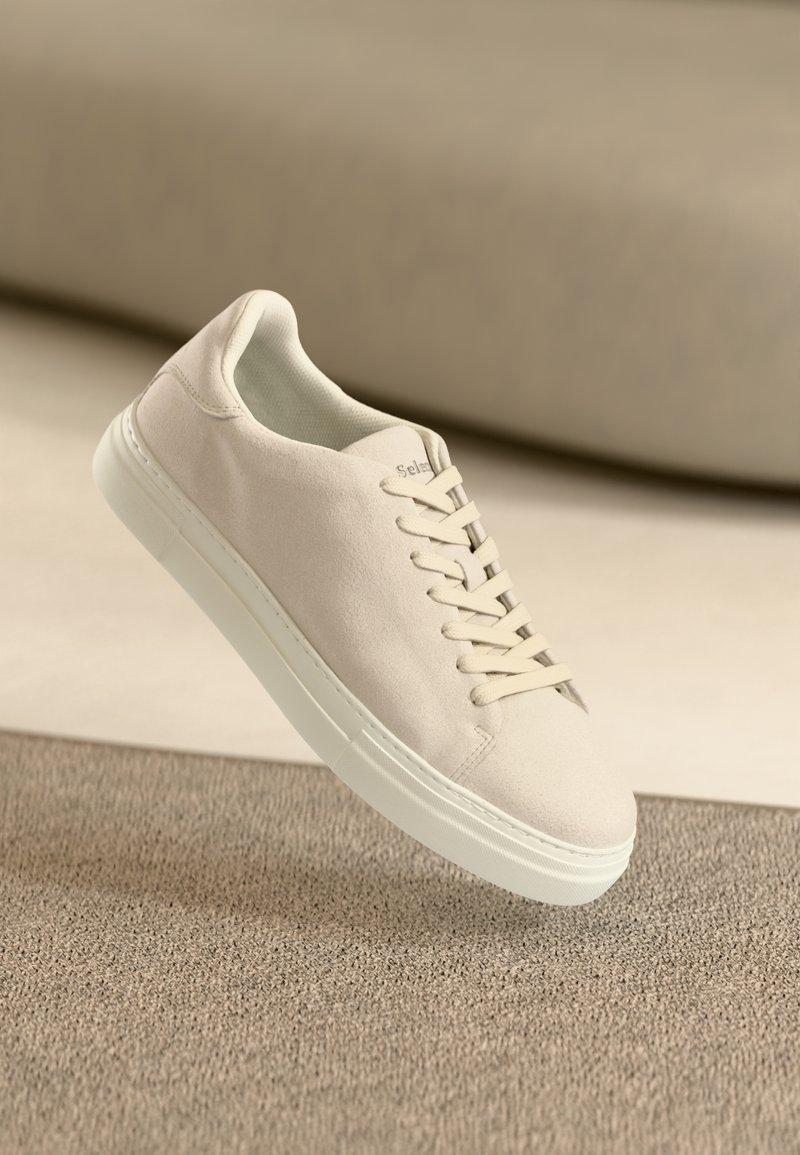 Baskets basses en daim beige clair avec semelle blanche et lacets, reposant sur une surface beige texturée sur un fond beige flou.