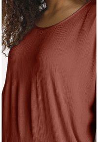 Rostfarbenes Oberteil mit einer strukturierten, zerknitterten Oberfläche. Hat einen weiten Ausschnitt und eine lockere Passform, die den fließenden Fall betont.