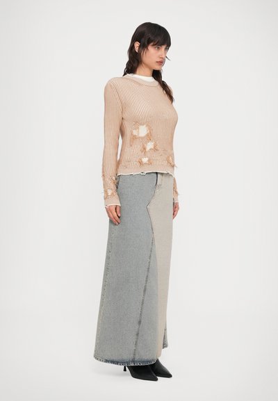 Femeie purtând un pulover bej din tricot deteriorat și o fustă lungă din denim bicolor, cu cizme negre cu vârf ascuțit, stând în picioare pe un fundal alb.