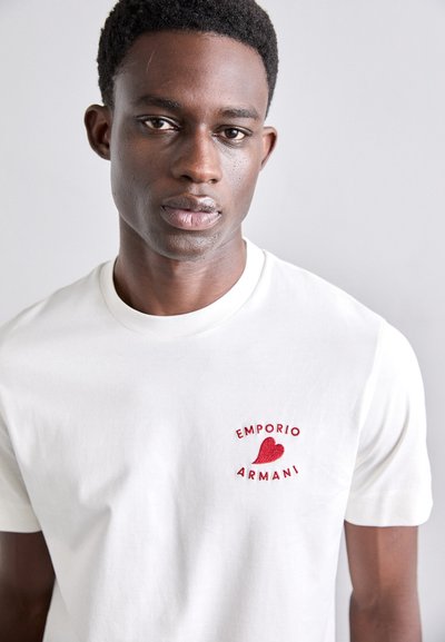 Valge puuvillane T-särk, millel on punane tikandiga logo "Emporio Armani" ja südamemuster vasakus rinnapiirkonnas. Sile tekstuur.