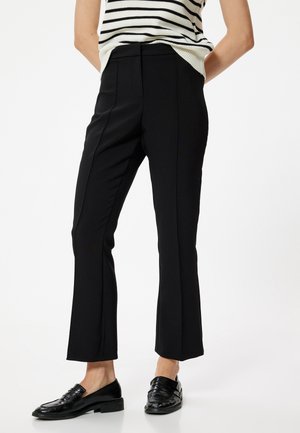 Pantalones negros de corte ajustado, con una tela suave y costuras frontales sutiles; combinados con mocasines negros brillantes.