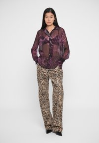 Translucent lilla bluse med bladmønster, knaplukning og lange ærmer; parret med beige leopardmønstrede bukser og sorte hæle.