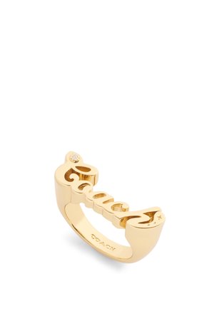 Gold ring featuring a cut-out script spelling "Coach," with a small gem accent on one end. Smooth, shiny finish with a rounded band.