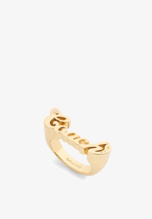 Gold ring featuring a cut-out script spelling "Coach," with a small gem accent on one end. Smooth, shiny finish with a rounded band.