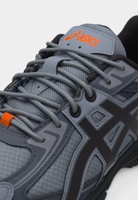 Κοντινό πλάνο γκρι και μαύρου παπουτσιού τρεξίματος ASICS με ύφασμα από δίχτυ, χοντρά κορδόνια και πορτοκαλί λογότυπο στη γλώσσα.