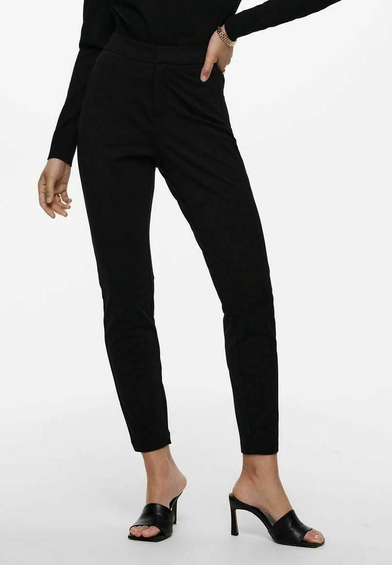 Pantalons noirs ajustés avec une coupe slim, présentant une texture lisse et une subtile couture avant. Associés avec des mules noires à talons.