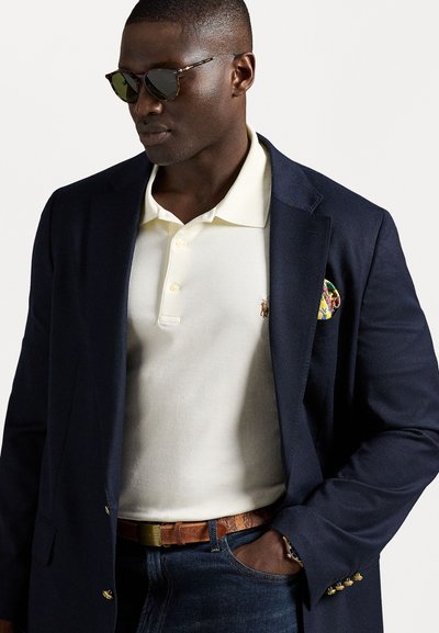 Blazer navy, polo galben deschis cu logo, blugi închiși, curea maro; ochelari de soare cu lentile întunecate și rame din acetat care imită țestoasa. Batistă cu un design colorat.