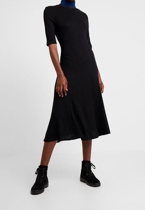 Vrouw die een zwarte midi-jurk draagt met ellebooglange mouwen, een blauwe coltrui en zwarte veterlaarzen tot aan de enkel, staand tegen een witte achtergrond.