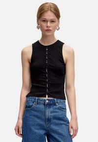 Zwarte mouwloze top gemaakt van ribbelmateriaal, met een knoopsluiting aan de voorkant en een cropped ontwerp. Gepaarde met highwaisted blauwe spijkerbroek.