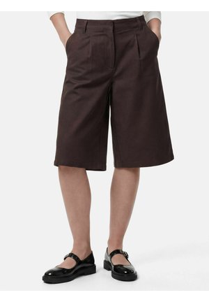 Brune knælange shorts med brede ben, båret med sorte Mary Jane-sko og en hvid top, hænderne i lommerne, stående på en hvid baggrund.